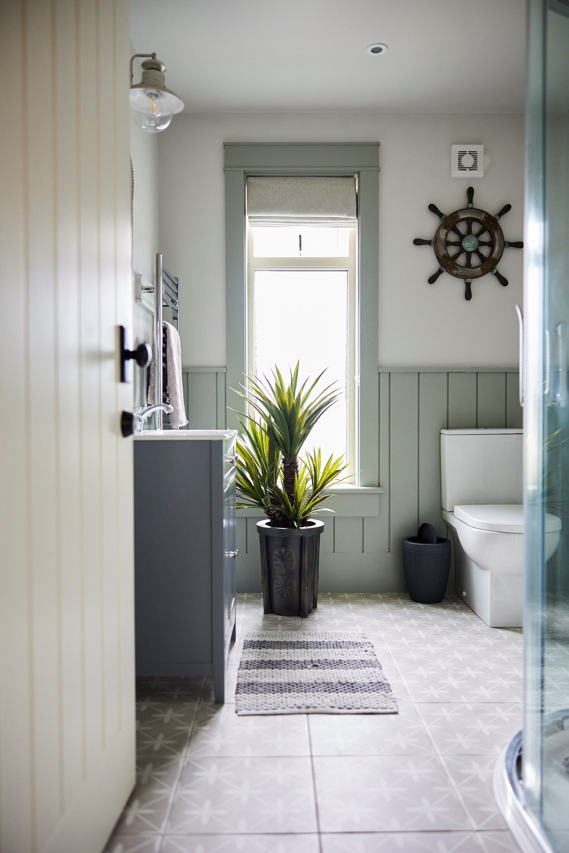 A bathroom with a toilet, a potted plant, a ship's wheel decoration, and a window with blinds.