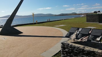 A coastal walkway with a metal sculpture, a grassy area, and a view of the sea and hills.