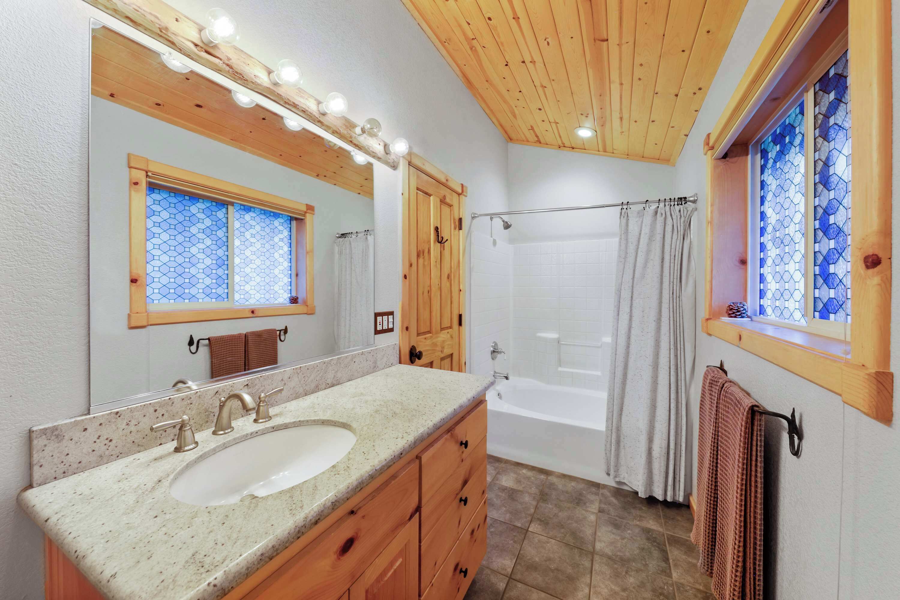 A bathroom with a wooden vanity, a large mirror with lights, a bathtub, and a window with stained glass.