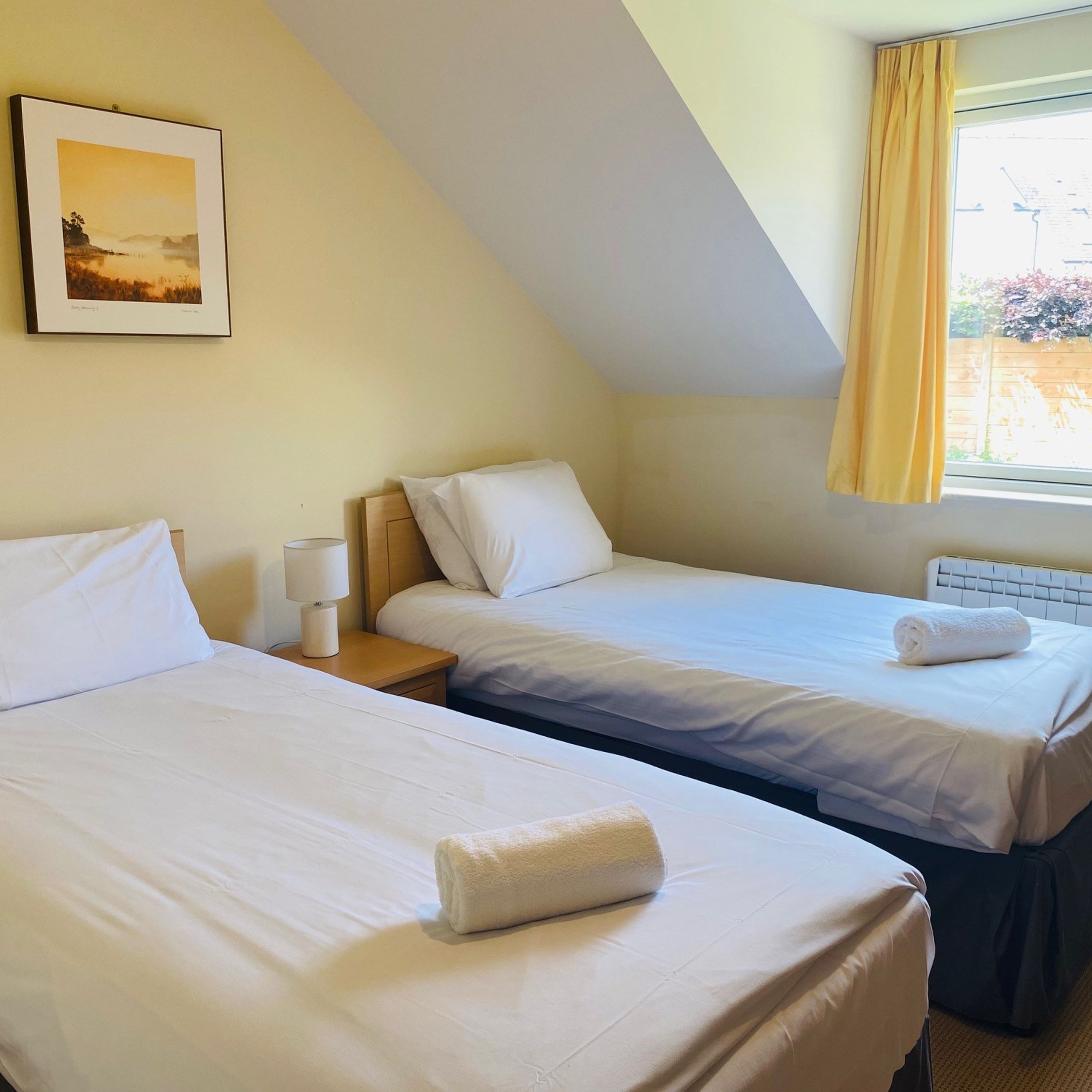 Een hotelkamer met twee enkele bedden, een nachtkastje met een lamp, een lijst met een afbeelding aan de muur en een raam met gordijnen.
