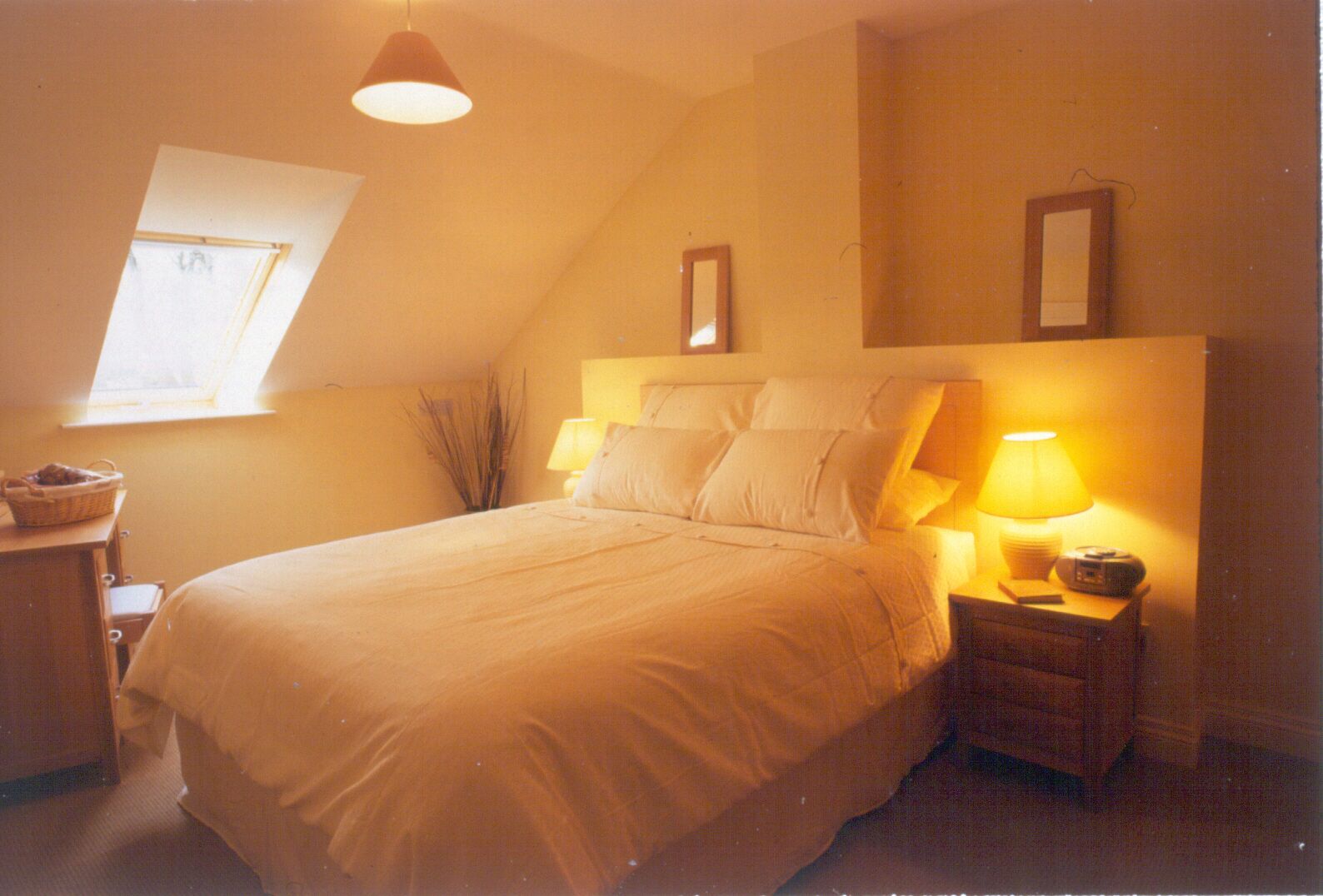 Une chambre avec un grand lit, deux tables de chevet avec des lampes, une petite table avec un panier, et une fenêtre donnant sur le ciel.