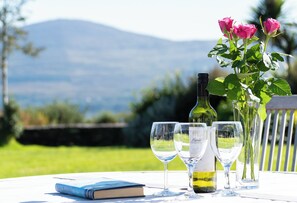 Una mesa con una botella de vino, copas y un libro, ubicada al aire libre con vistas a la montaña.