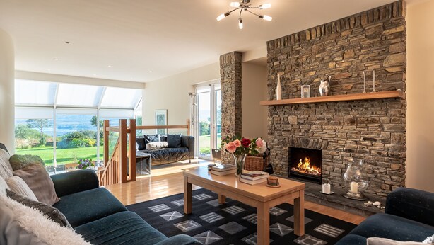 A modern living room with a stone fireplace, a wooden coffee table, and a view of the outdoors. - A Split-level Spacious Home With sea Mountain Views (Greenane)