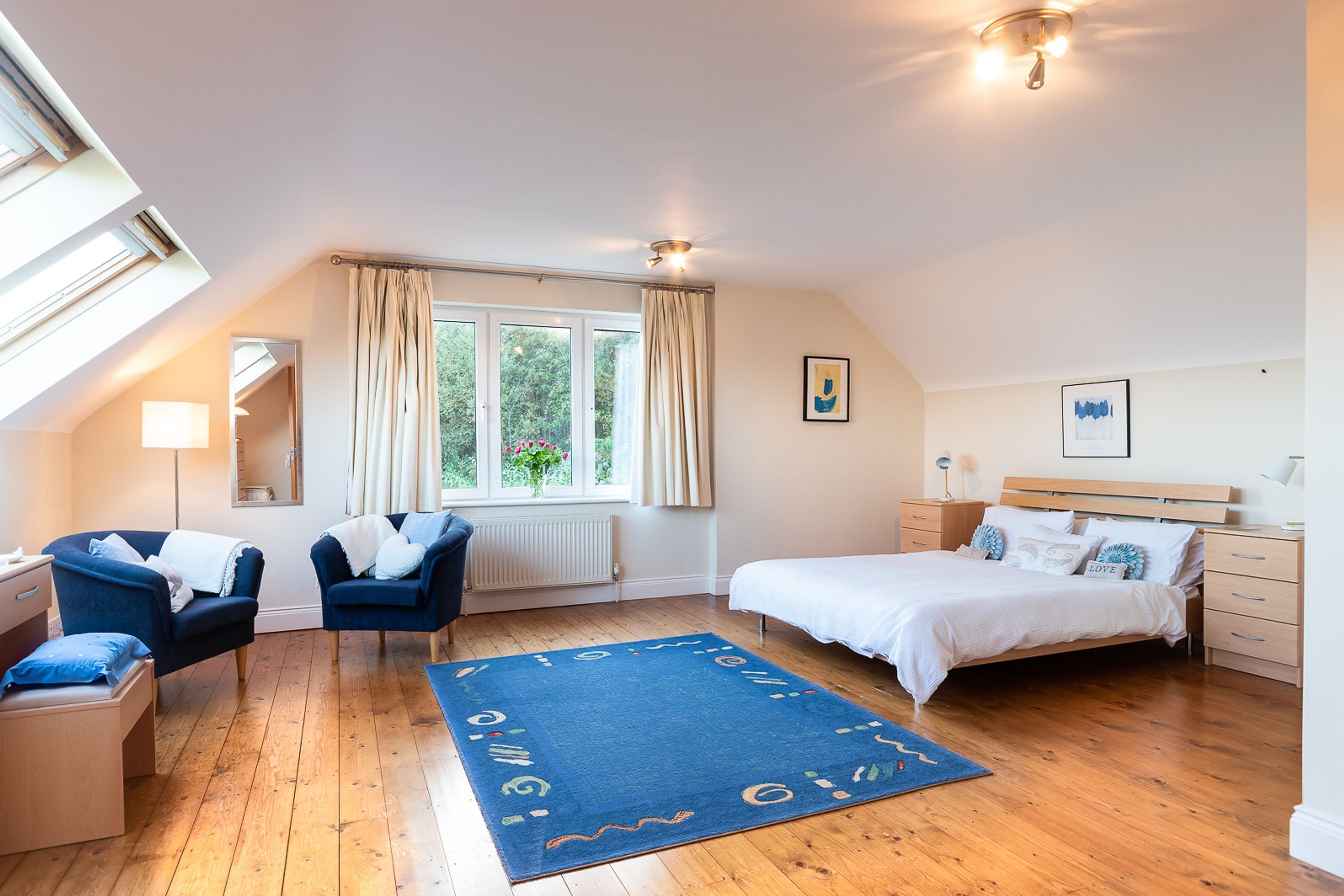 A bedroom with a sloped ceiling, a large window with curtains, a bed with white bedding, a blue armchair, a bedside table, and a blue rug.