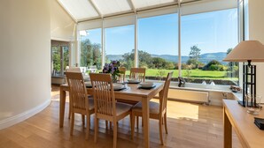 A dining area with a table set for four, large windows offering a view of a green landscape, and a lamp on the table. - A Split-level Spacious Home With sea Mountain Views (Greenane)