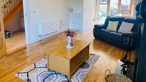 A spacious living room with a wooden floor, a black sofa, a wooden coffee table, and a vase of flowers. - Holiday Home Within Easy Walking Distance to Kenmare (Kenmare)
