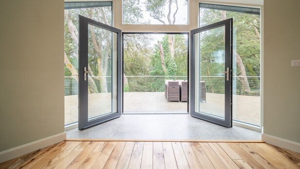 Une pièce dotée d’un plancher en bois, de grandes portes vitrées donnant sur un espace extérieur avec vue sur les arbres et un jardin clos.