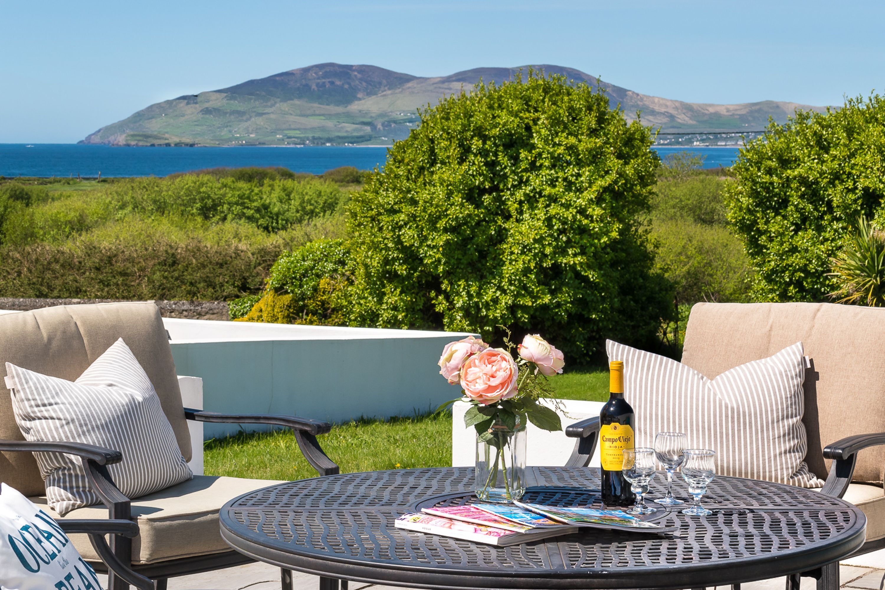 A round metal table with a bottle of wine, glasses, and a book. There are two cushioned chairs with pillows. A vase with flowers is on the table. The background features a view of the sea and hills.