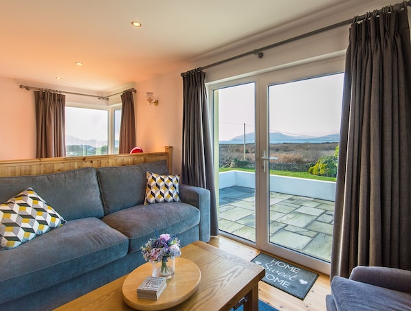 A modern living room with a sofa, a coffee table, and a view of a scenic landscape through large sliding glass doors. - Large House With Adjoining Cottage Close to Waterville (Waterville)
