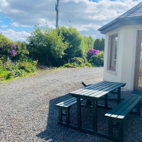 Un sendero de grava conduce a una mesa de picnic de madera con bancos, rodeado de abundante follaje y flores moradas.