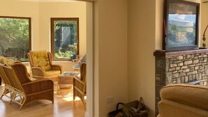 A spacious living room with a stone fireplace, wicker furniture, and large windows offering a view of greenery. - 3 Bedroom Home 15 Mins Drive From Kenmare Town (Dawros)