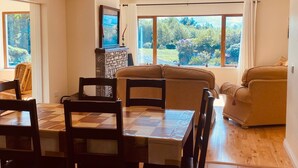 A dining area with a wooden table and chairs, a stone fireplace, and large windows offering a view of the outdoors. - 3 Bedroom Home 15 Mins Drive From Kenmare Town (Dawros)