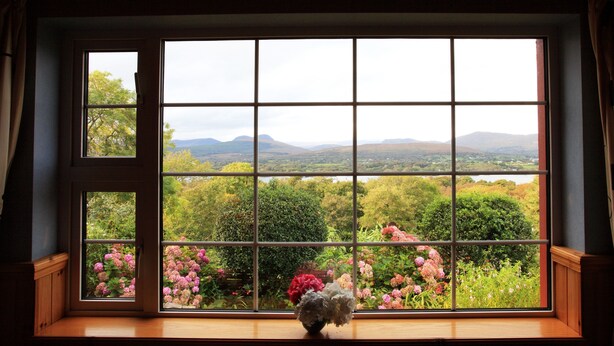 A room with a large window offering a view of a lush garden and distant mountains. - 3 bed Seaview Cottage (Dawros)