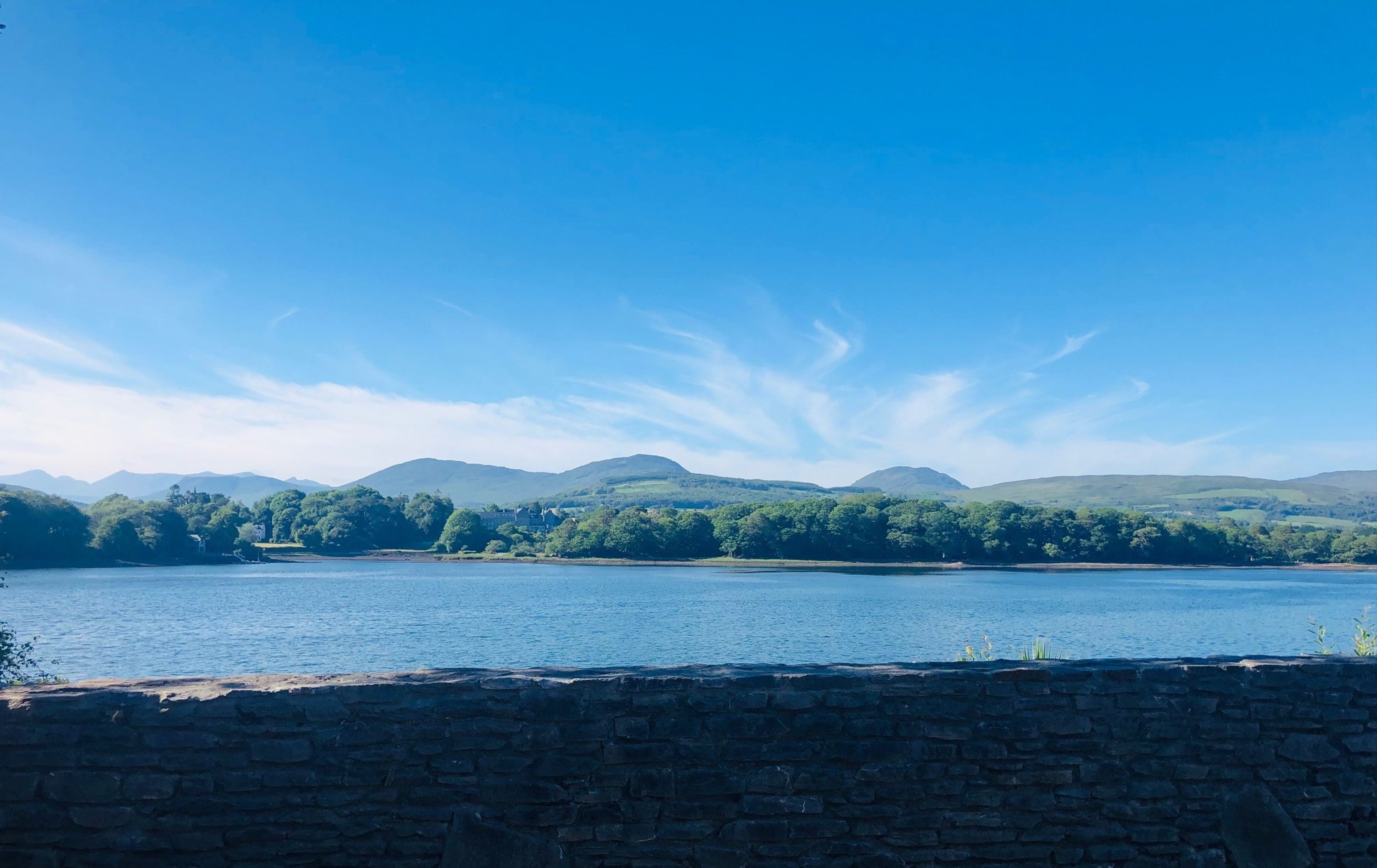 A stone wall and a body of water with distant mountains and trees.