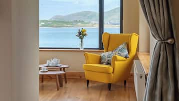 A room with a yellow armchair by a large window overlooking a body of water and hills. There's a small table with a teapot and cups, and a vase with flowers on the windowsill.