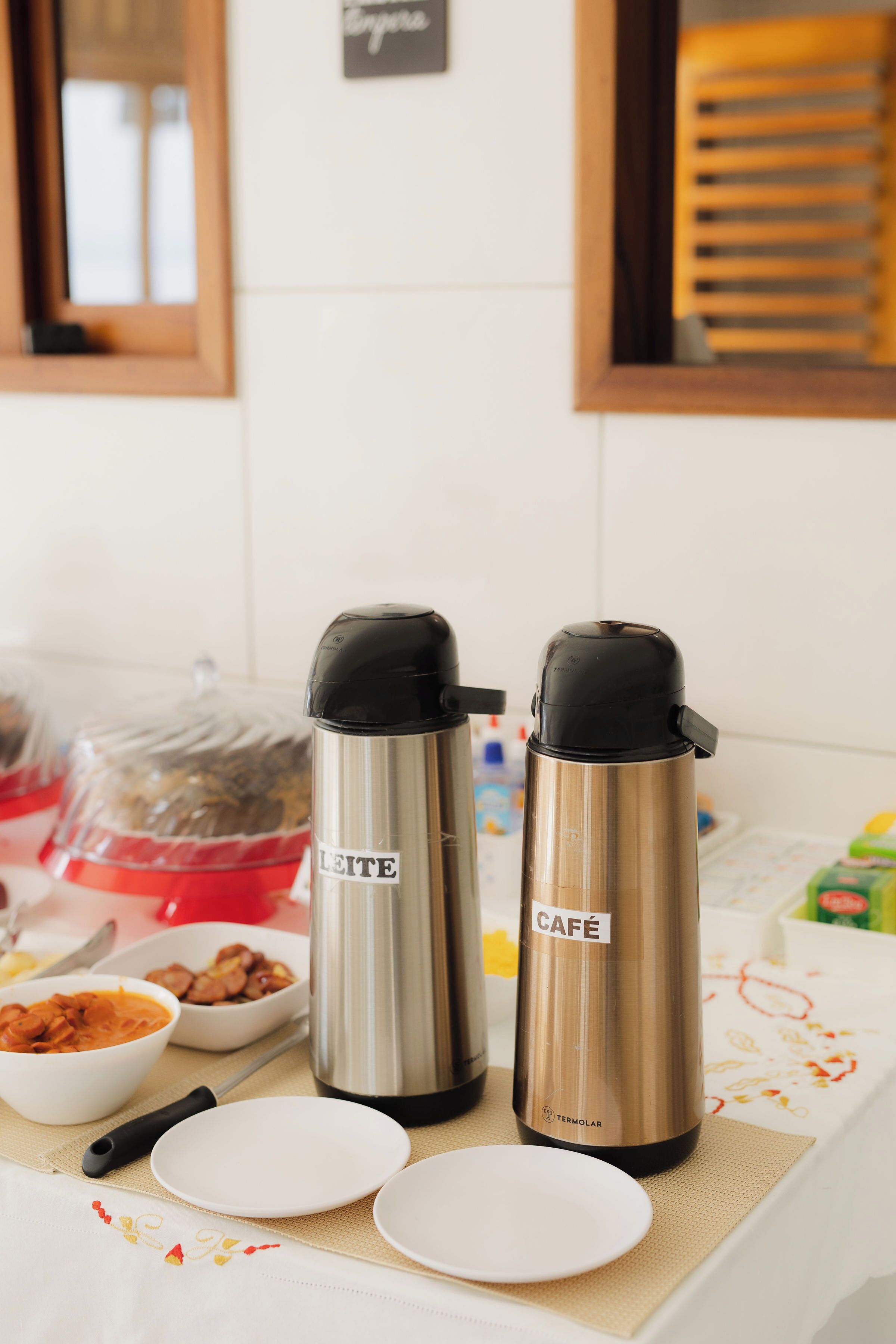 A table with two stainless steel thermos cups labeled "LEITE" and "CAFÉ" and a few bowls of food.