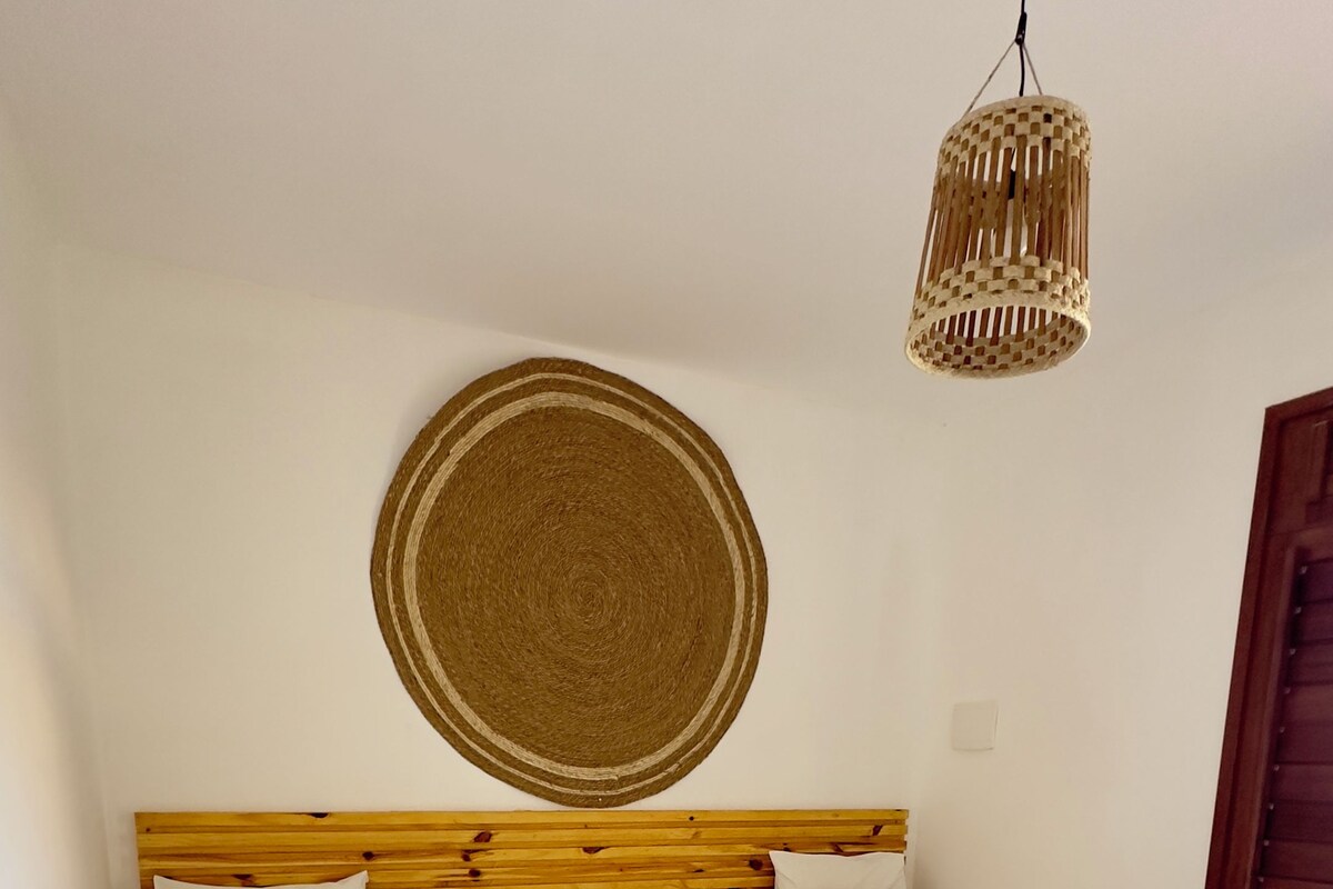 Una habitación con dos camas, un tapiz de pared tejido, una mesita de noche pequeña y blanca, y una lámpara colgante.