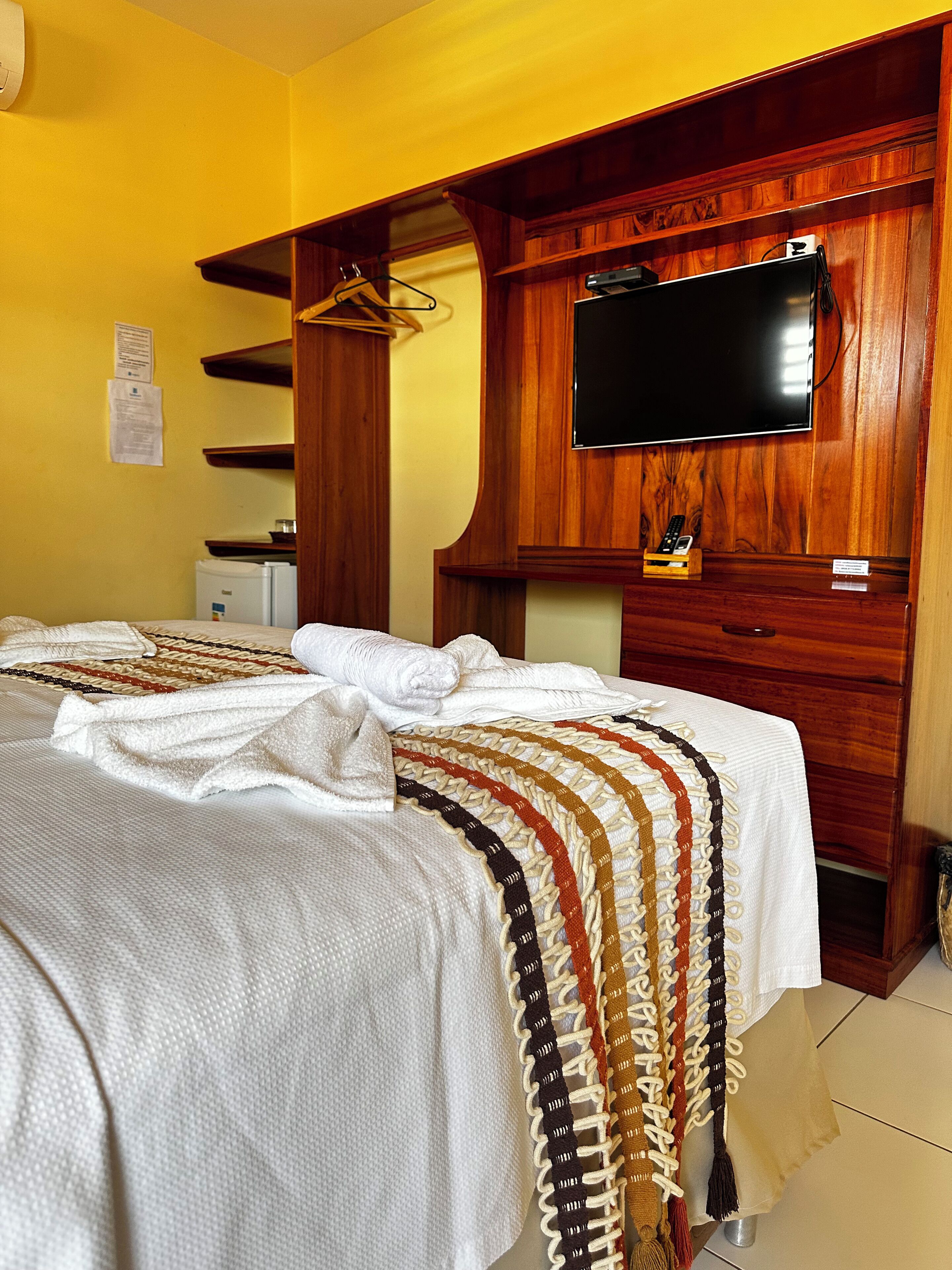 A hotel room with a bed, a television mounted on a wooden wall, a small refrigerator, and a wooden cabinet.