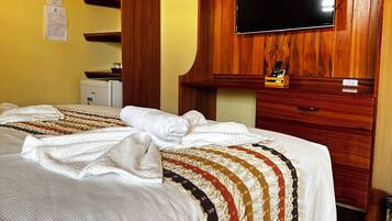 A hotel room with a bed, a television mounted on a wooden wall, a small refrigerator, and a wooden cabinet.