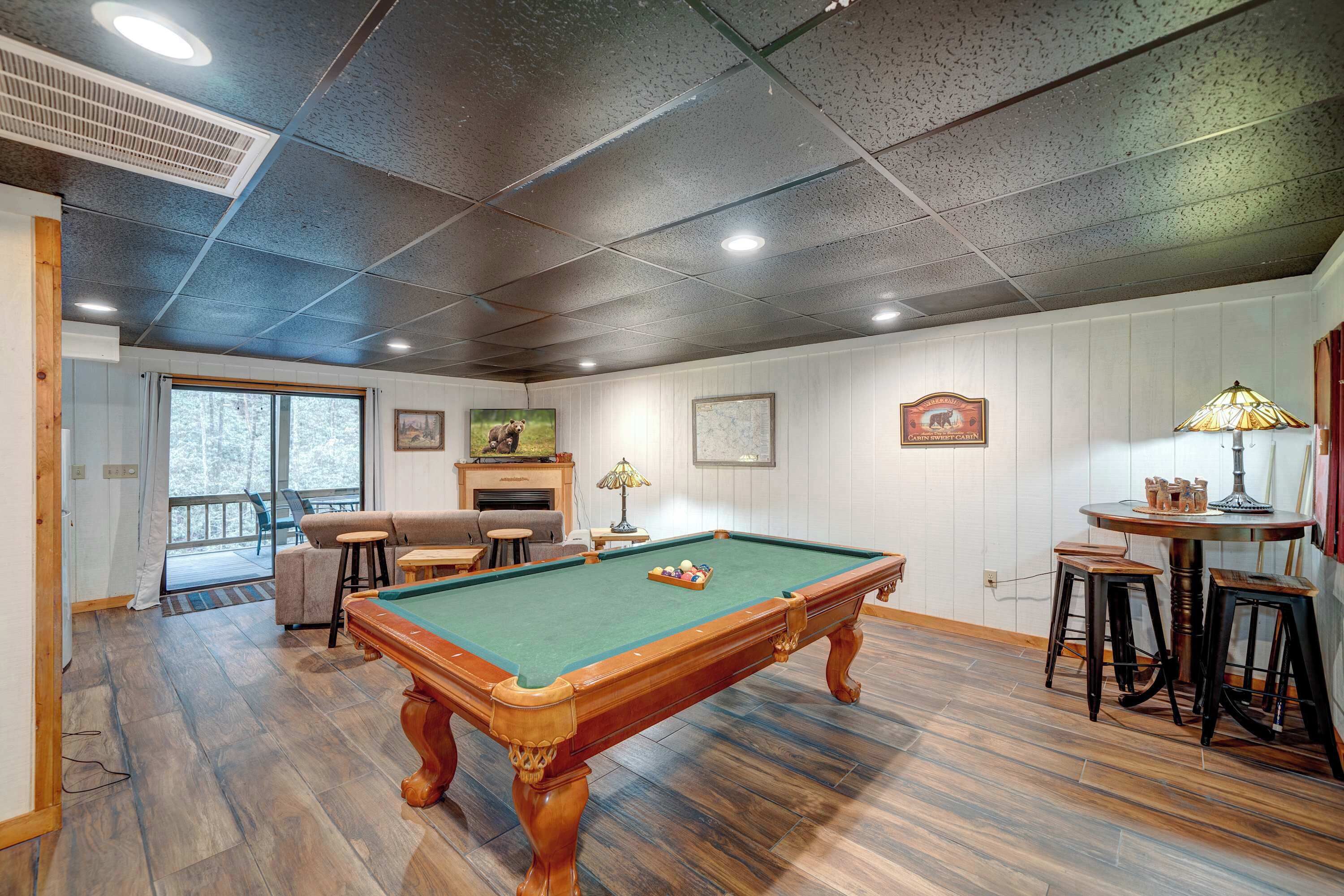 A pool table in a room with wooden flooring, a fireplace, and a dining area with stools.