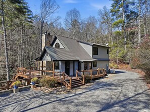 A two-story cabin with a stone chimney, wooden deck, and a gravel driveway.