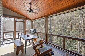 A wooden deck with a table and chairs, a ceiling fan, and a view of a forest.