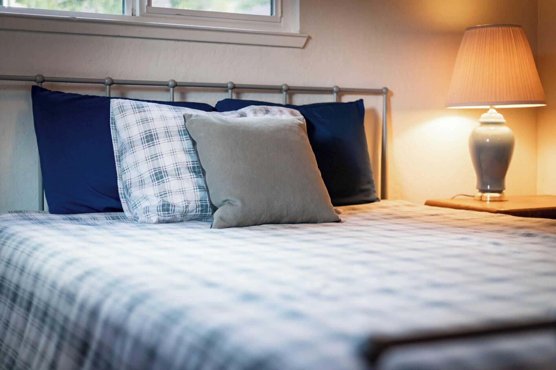 A neatly made bed with a blue and white checkered bedspread, a grey pillow, and two navy blue pillows. A bedside lamp with a beige shade is on a wooden nightstand to the right.