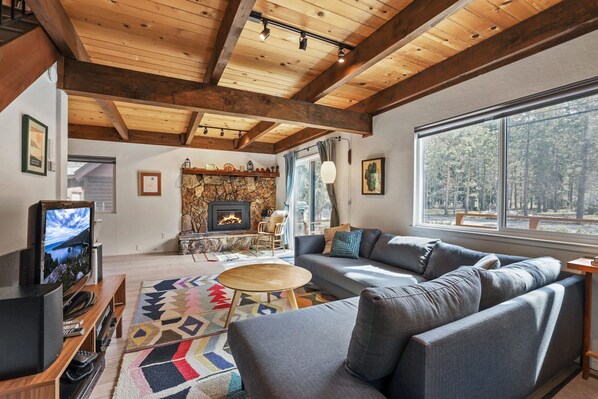 Ein gemütliches Wohnzimmer mit Kamin aus Stein, einem großen Fenster mit Blick auf den Wald und einem modernen Sofa.