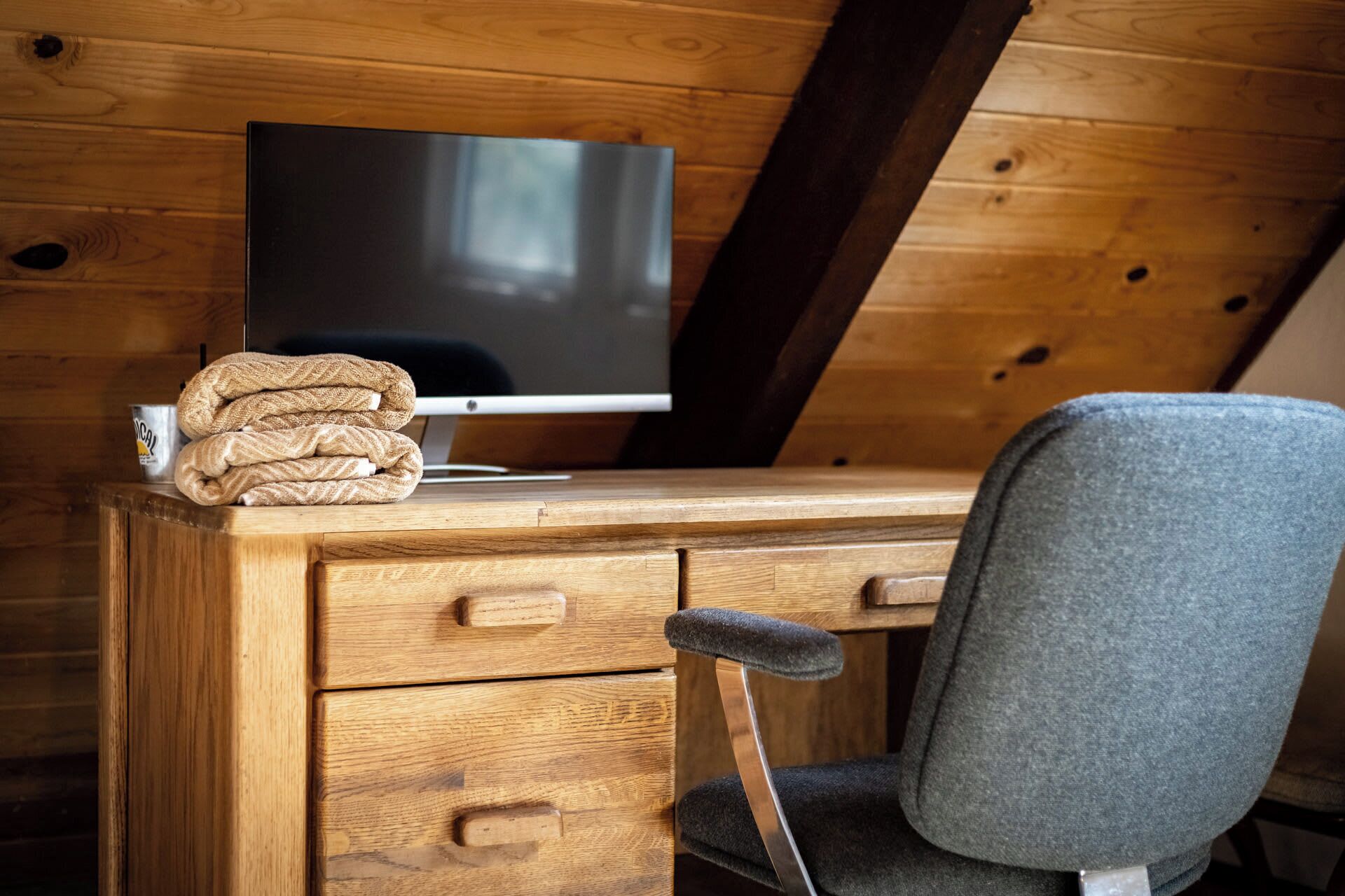 A wooden desk with a computer monitor, a stack of towels, and a grey office chair in a room with wooden walls.