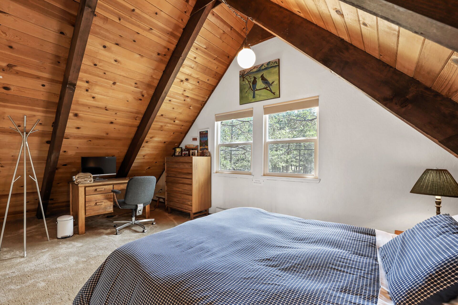 A cozy attic bedroom with a sloped ceiling, wooden beams, a desk with a chair, a bed with a patterned bedspread, and two windows offering a view of trees.