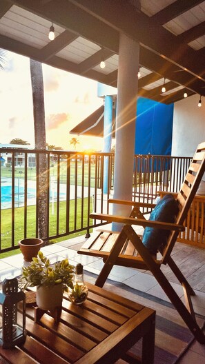 A wooden lounge chair with a cushion, a small table with a potted plant, and a railing overlooking a pool area.