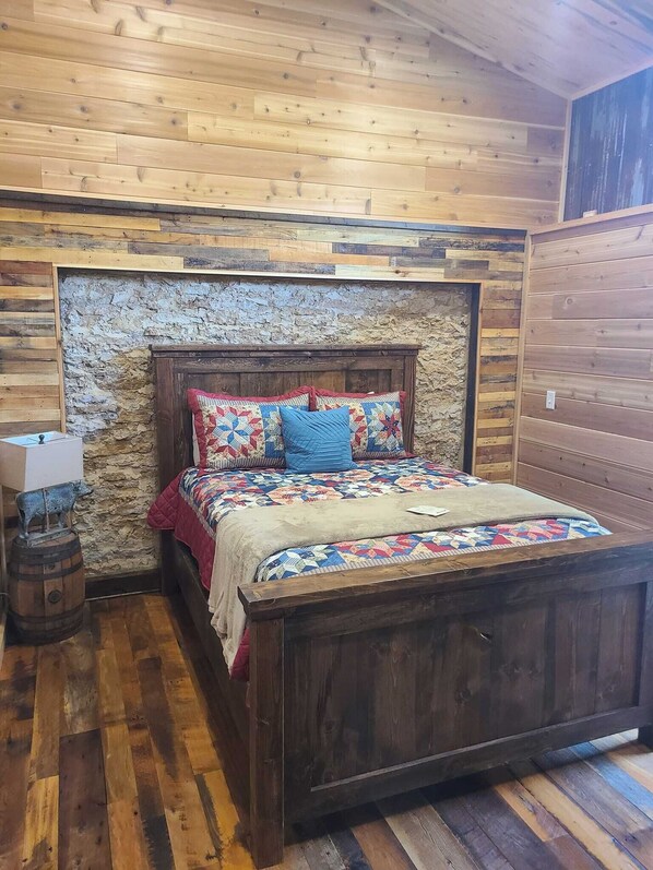 Una cama de madera con colcha acolchada y almohadas, una pared de piedra rústica y un suelo de madera.