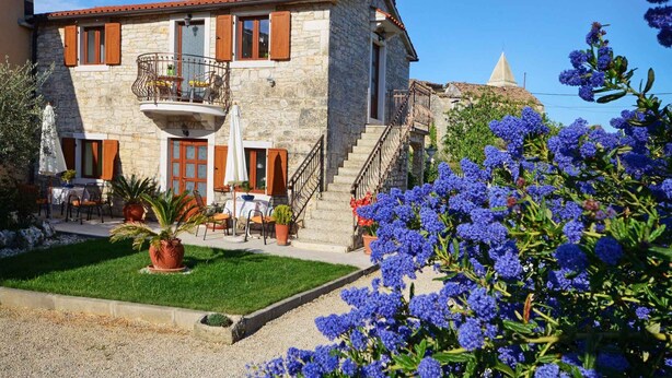 A stone building with a balcony, outdoor seating, and a garden with blue flowers.