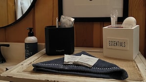 Un baño con un cuadro de un ciervo enmarcado, un perchero con una toalla, una botella negra de sal marina, un recipiente negro, una caja de pañuelos y una caja con la etiqueta 'ESSENTIALS'.