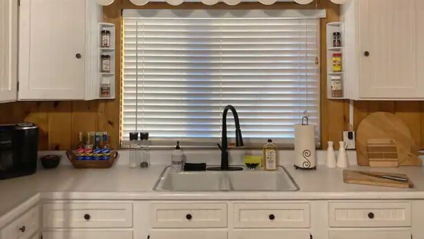 Una cocina con armarios blancos, un grifo negro y una ventana con persianas.