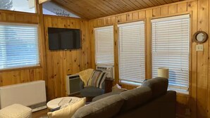 Interior acogedor de una cabaña con paredes de madera, un sofá, una silla, un televisor y una ventana con persianas.