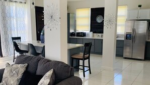 A modern living room with a gray sofa, patterned pillows, and a geometric rug. A dining area with a table and chairs is visible in the background.