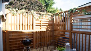 Une terrasse en bois avec un barbecue, deux chaises, une table ornée d’une plante en pot et une bouteille d’eau.