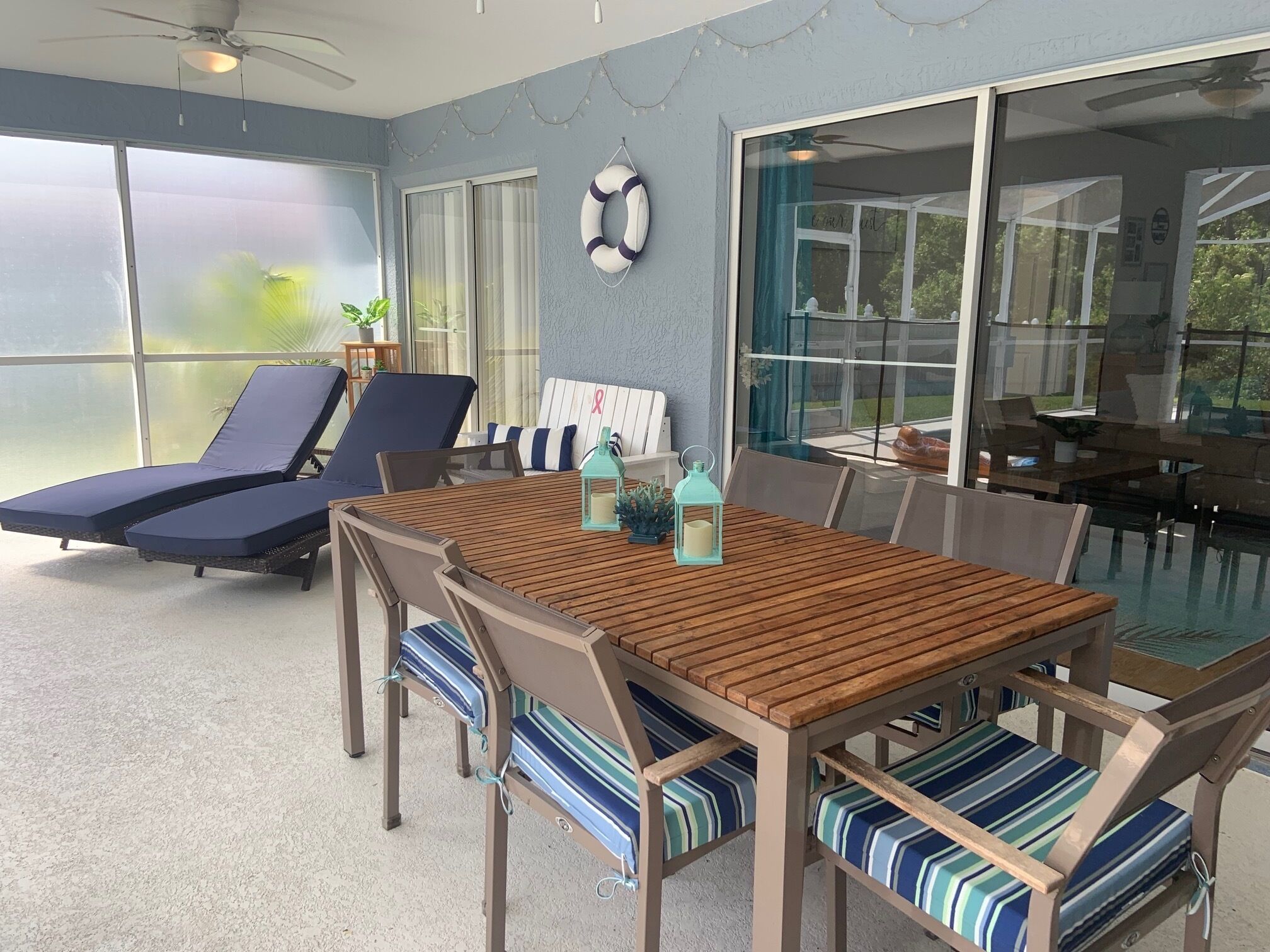 A sunlit patio with a wooden table and striped cushioned chairs, a ceiling fan, and a large sliding glass door leading to a garden.