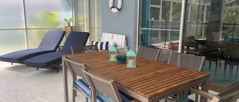 A sunlit patio with a wooden table and striped cushioned chairs, a ceiling fan, and a large sliding glass door leading to a garden.