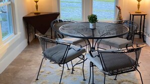 Un coin repas comprenant une table ronde et quatre chaises, un ventilateur de plafond et deux fenêtres donnant sur un paysage verdoyant.