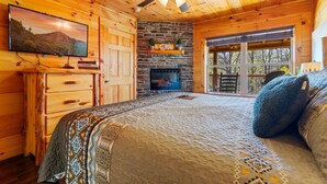 A cozy log cabin bedroom with a fireplace, a television, and a large bed with patterned bedding.