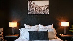 A bed with striped bedding, two bedside tables with lamps, a black and white leopard print artwork on the wall, and a small plant on the right side. - Home In The Heart Of Alvin! (Alvin)