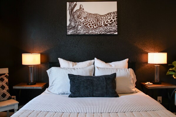 A bed with striped bedding, two bedside tables with lamps, a black and white leopard print artwork on the wall, and a small plant on the right side.