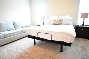 A bedroom with a bed, a nightstand, a lamp, a sofa, and a window.
