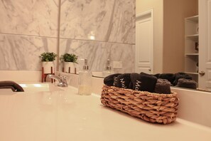 A bathroom with a marble wall, a sink, a mirror, a towel basket, and a door.
