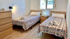 Une chambre avec deux lits, une commode en bois, une table de chevet avec une lampe et une fenêtre donnant sur l’extérieur.