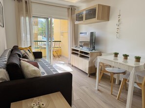 A modern living room with a sofa, a dining table with chairs, a TV, and a balcony with a view. - On the Beach Holiday Home (Los Cristianos)