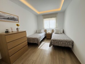 A bedroom with two beds, a wooden dresser, a window with blinds, and a ceiling light fixture. - On the Beach Holiday Home (Los Cristianos)