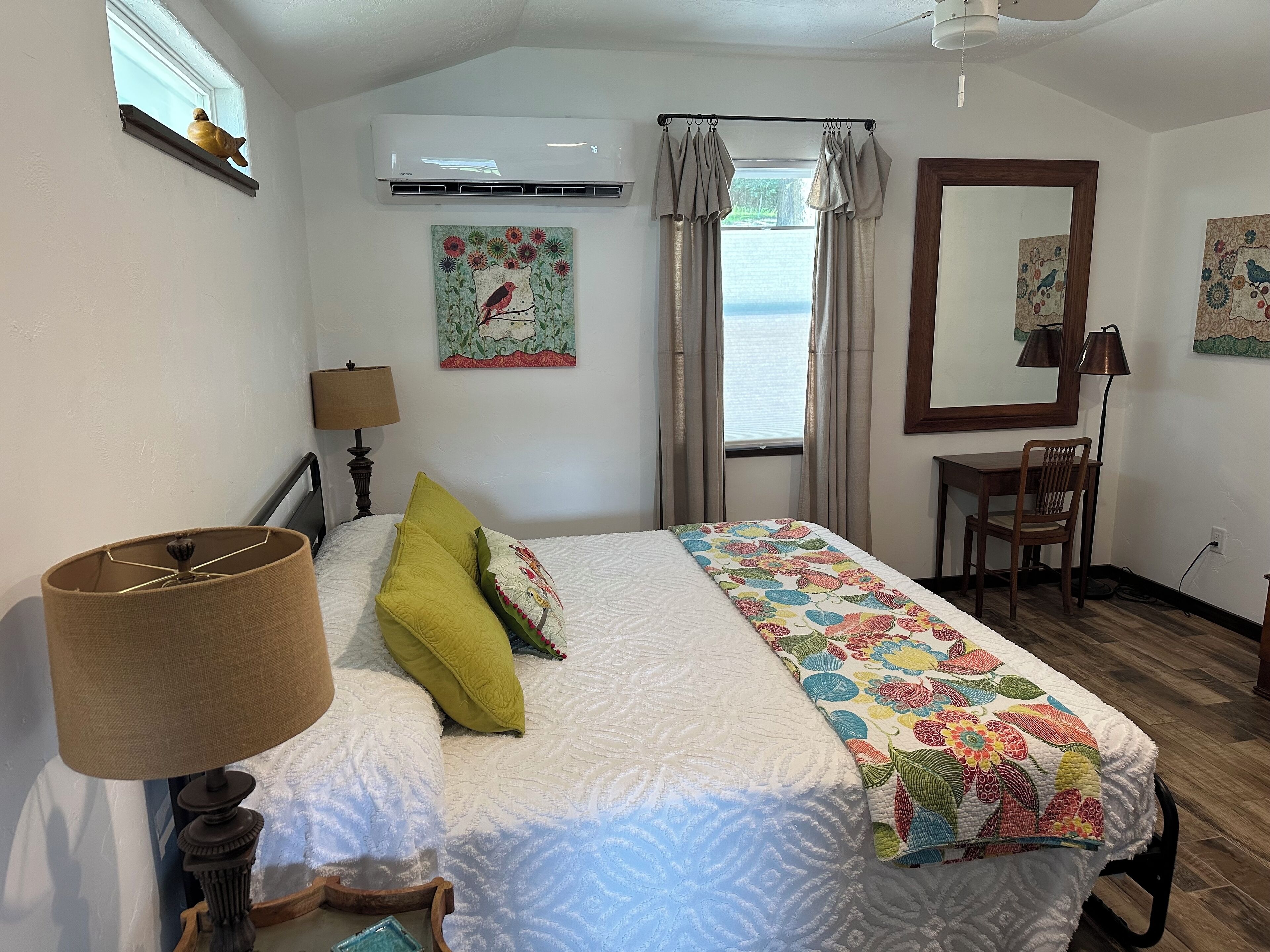 A bedroom with a bed, a desk, a chair, an air conditioner, a window with curtains, and a wall with a painting.